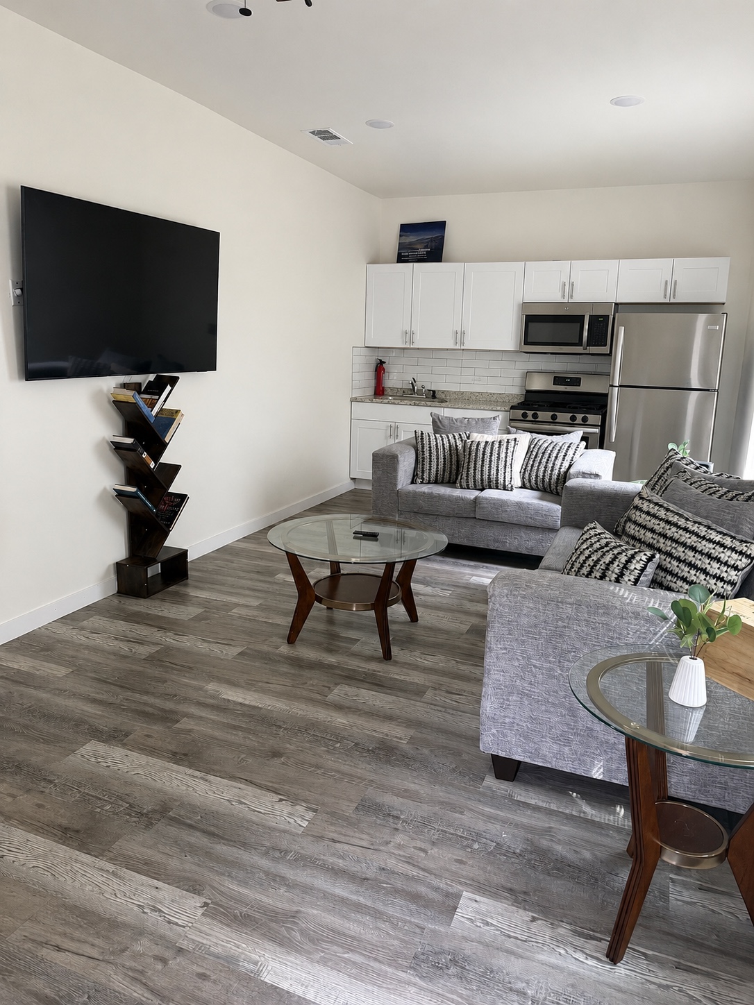 Living room with grey sofas, glass coffee table, wall-mounted TV, and a tree-shaped bookshelf.